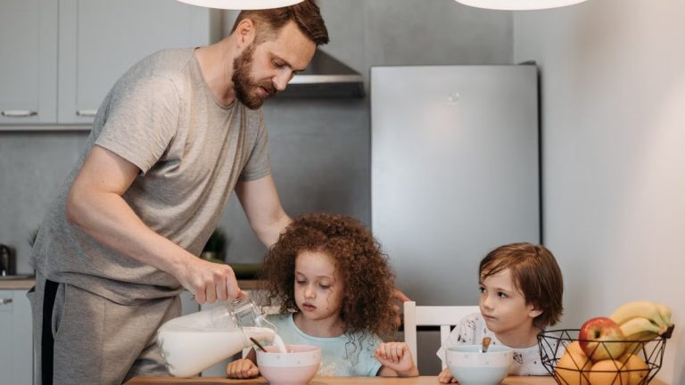 A father pours milk into cereal bowls for his children at the breakfast table in a modern kitchen.
